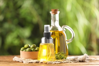 Photo of Bottles with cooking oil, olives and rosemary on wooden table against blurred background