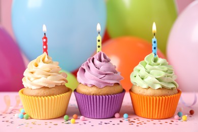 Photo of Birthday cupcakes with burning candles and sprinkles on pink table against color balloons, closeup