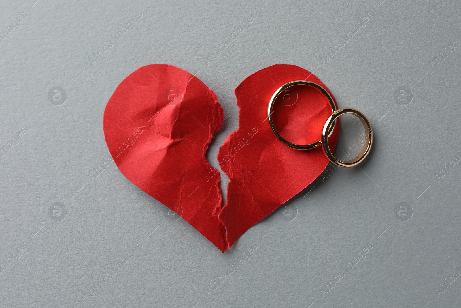 Photo of Halves of torn red paper heart and wedding rings on white background, top view. Broken heart