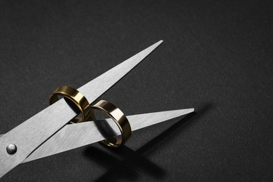 Photo of Divorce concept. Scissors with wedding rings on blades against black background, closeup. Space for text