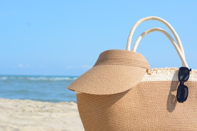 Photo of Stylish straw bag with visor cap and sunglasses on beach near sea, closeup. Space for text