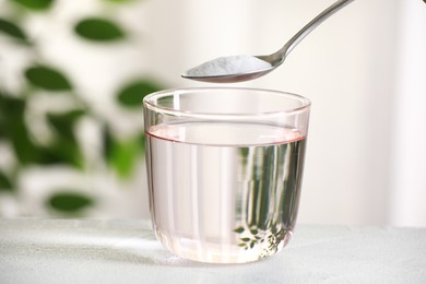 Spoon with baking soda over glass of water at white table against blurred background, closeup