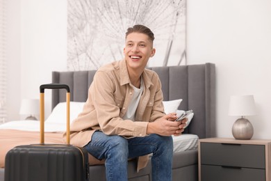 Photo of Smiling guest with suitcase and smartphone on bed in stylish hotel room