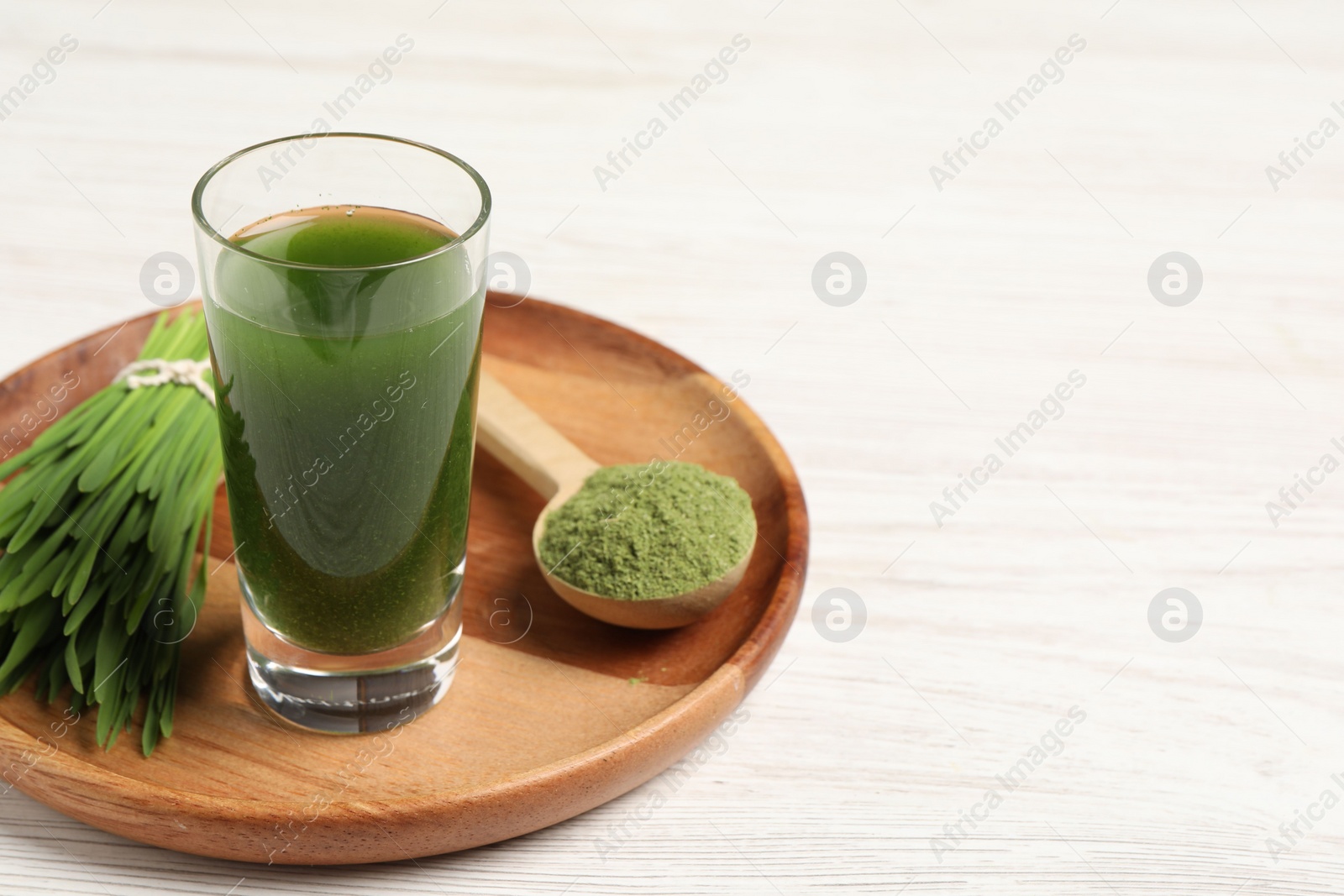 Photo of Wheat grass drink in shot glass, spoon of green powder and fresh sprouts on white wooden table, space for text