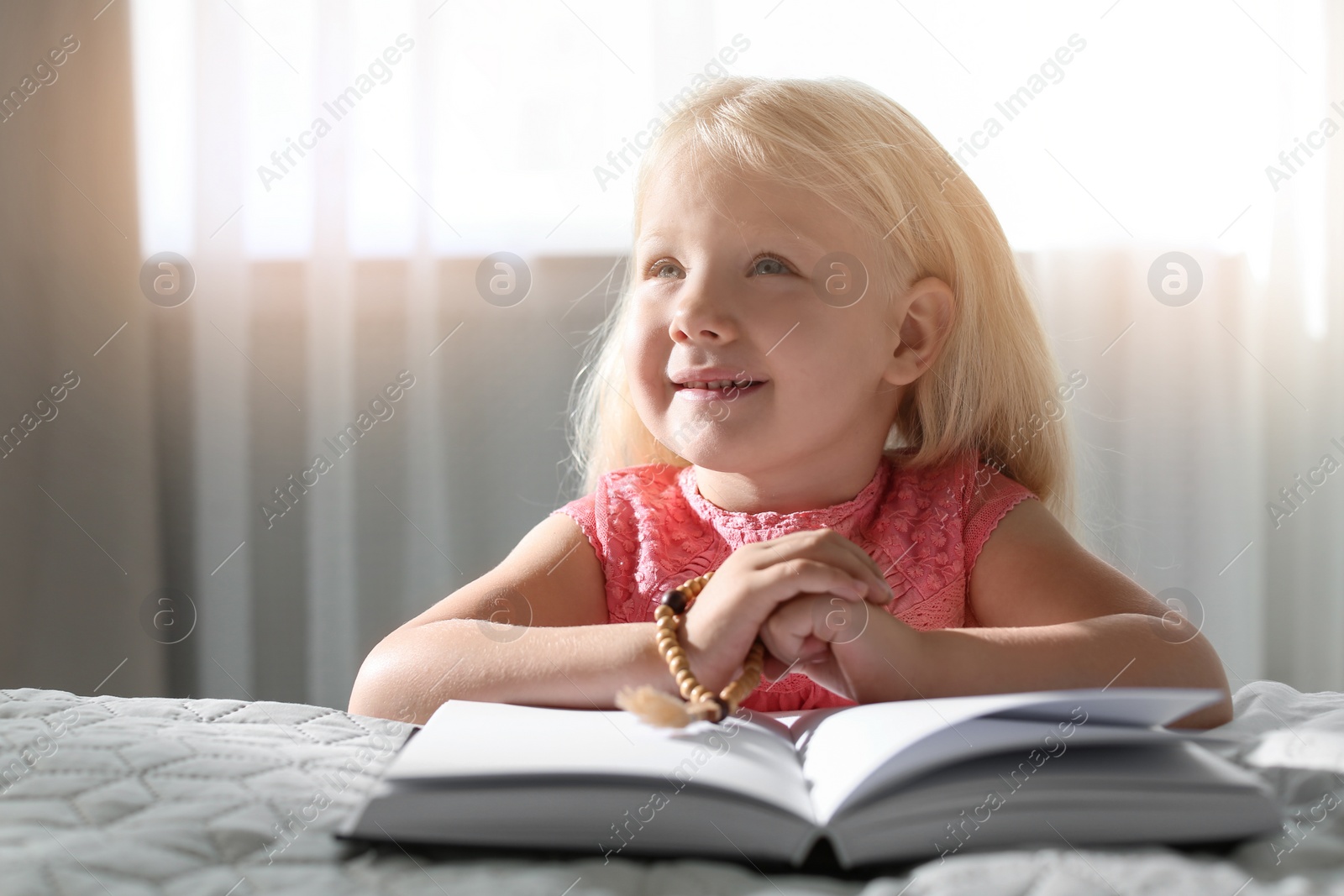 Photo of Cute little girl with beads praying over Bible in bedroom