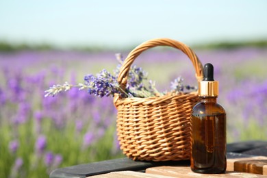 Bottle of essential oil and wicker bag with lavender flowers on wooden table in field outdoors, space for text