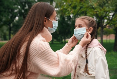 Mother putting mask onto face of little daughter in park during coronavirus pandemic