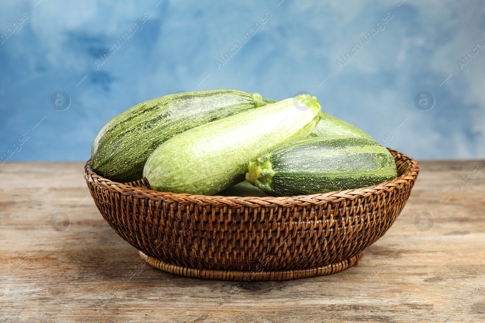 Photo of Wicker bowl with fresh ripe green zucchini on wooden table against blue background