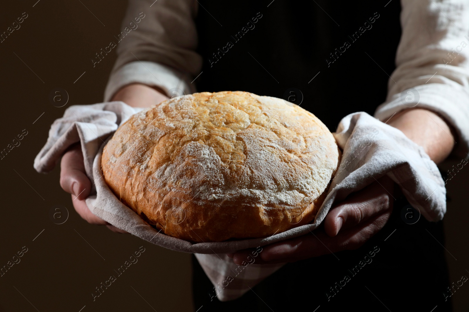 Photo of Man holding loaf of fresh bread on dark brown background, closeup