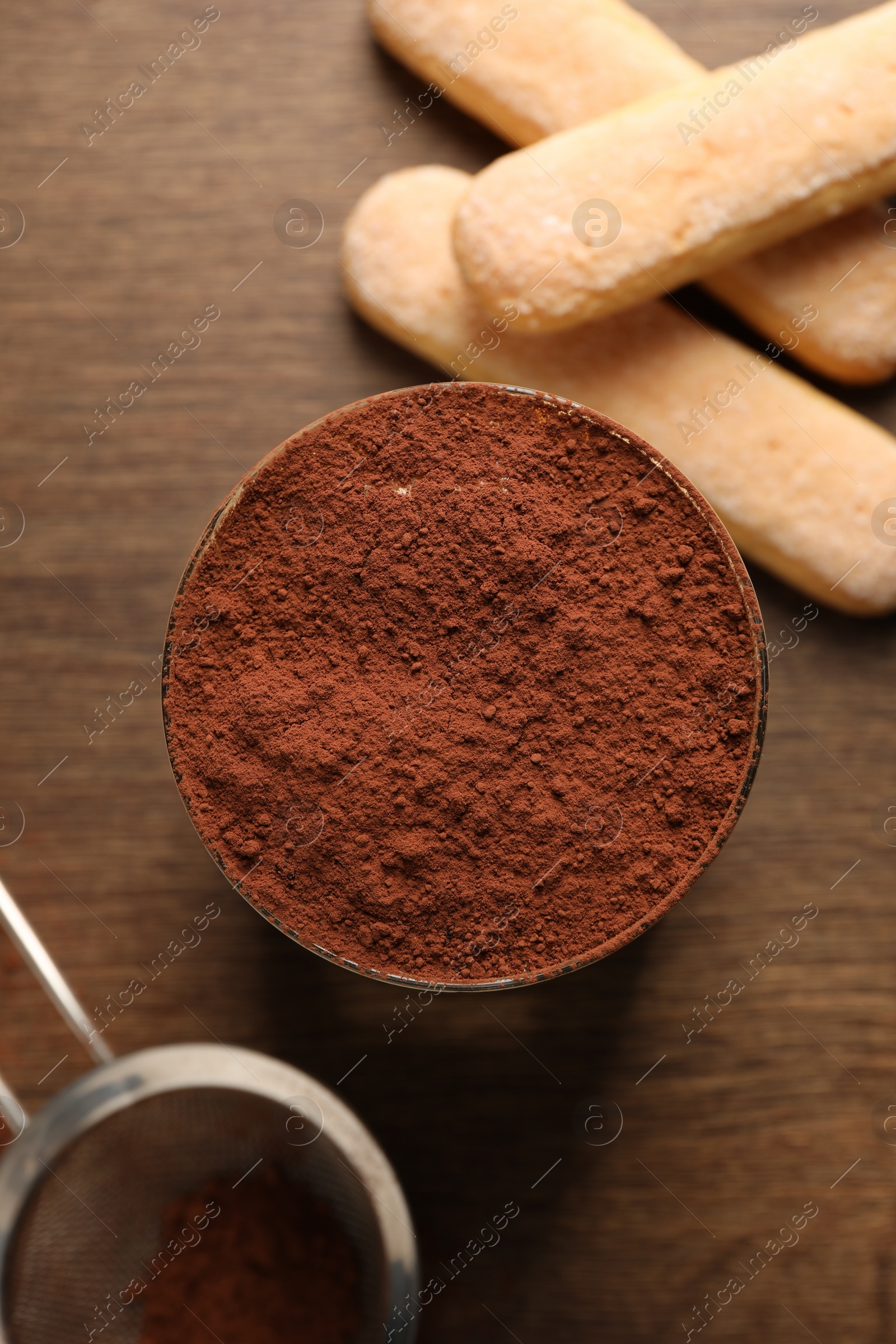 Photo of Delicious tiramisu in glass, biscuits and sieve with cocoa powder on wooden table, flat lay