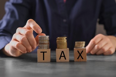 Photo of Woman with word Tax made of wooden cubes and coins at grey table, closeup