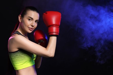 Photo of Portrait of beautiful woman wearing boxing gloves in color lights and smoke on black background. Space for text
