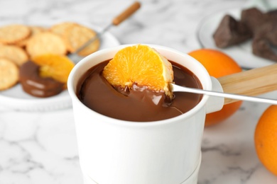 Dipping orange into fondue pot with chocolate on white marble table, closeup