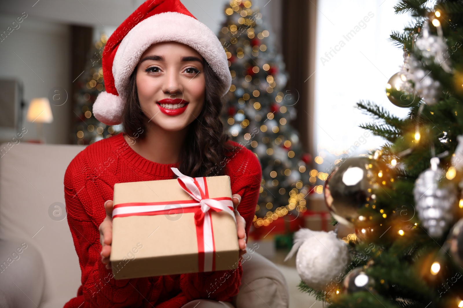 Photo of Beautiful woman in Santa hat with gift box near Christmas tree at home