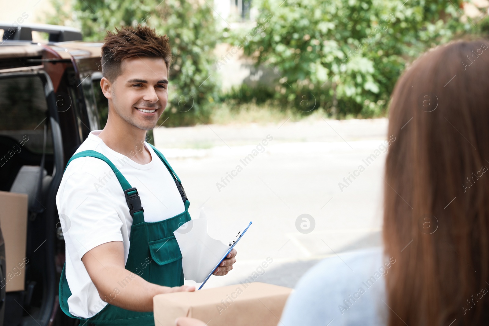 Photo of Young delivery courier with clipboard giving parcel to customer outdoors