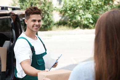 Young delivery courier with clipboard giving parcel to customer outdoors