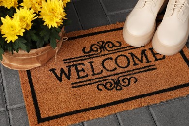 Photo of Door mat with word Welcome, stylish boots and beautiful flowers on floor