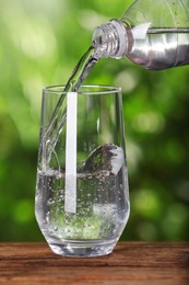Photo of Pouring water from bottle into glass on wooden table outdoors, closeup