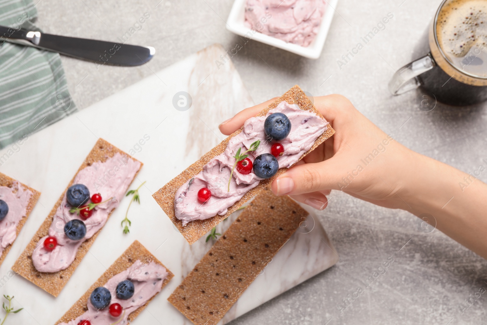 Photo of Woman holding tasty cracker sandwich with cream cheese, blueberries, red currants and thyme at grey table, top view