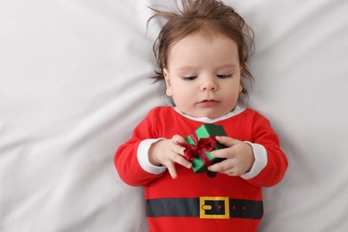 Cute baby wearing festive Christmas costume with gift box on white bedsheet, top view
