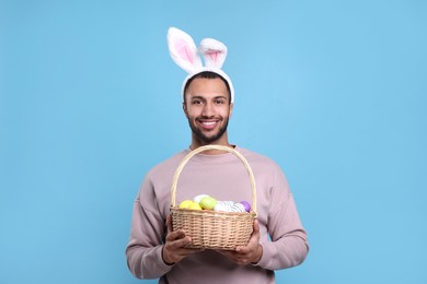 Photo of Happy African American man in bunny ears headband holding wicker basket with Easter eggs on light blue background