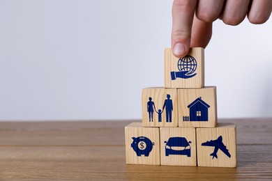 Image of Man building pyramid of cubes with different icons on wooden table against light background, closeup. Insurance concept