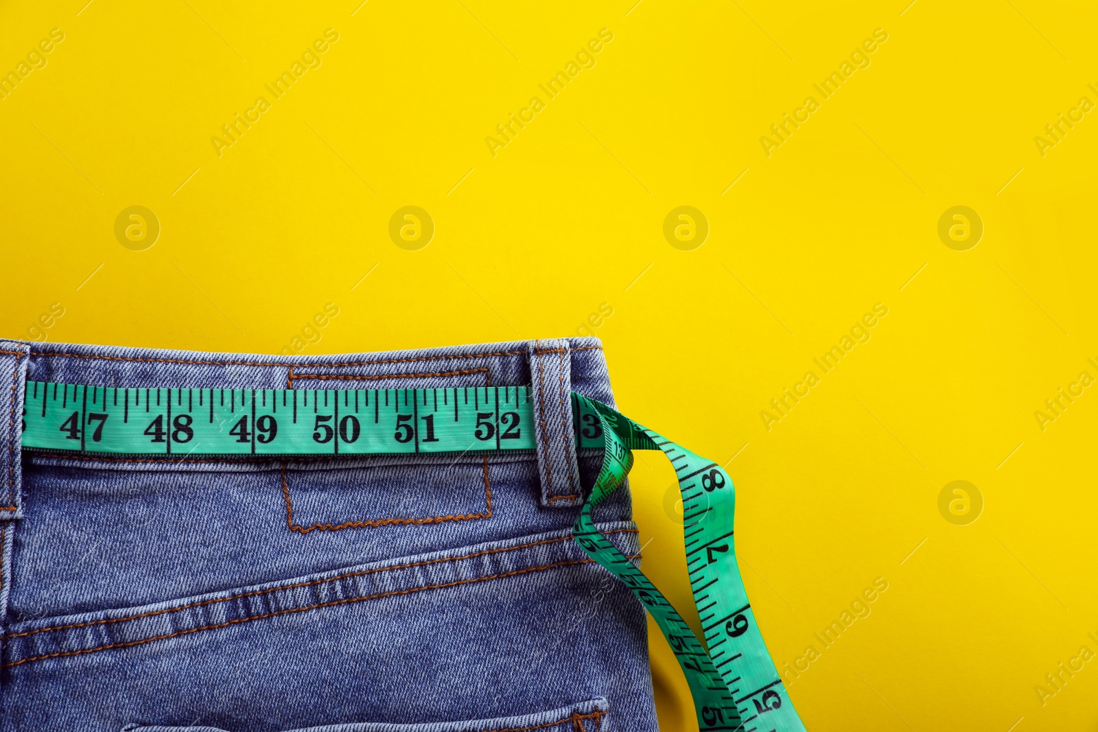 Photo of Jeans with measuring tape on yellow background, top view and space for text. Weight loss concept