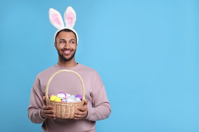 Happy African American man in bunny ears headband holding wicker basket with Easter eggs on light blue background, space for text