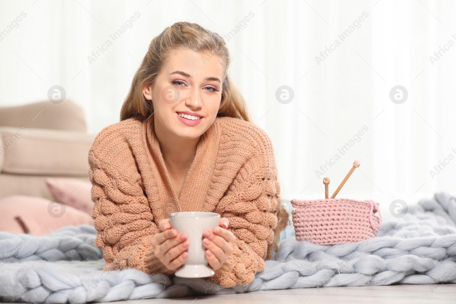 Photo of Attractive smiling young woman in cozy warm sweater with cup of hot drink lying on floor at home. Space for text