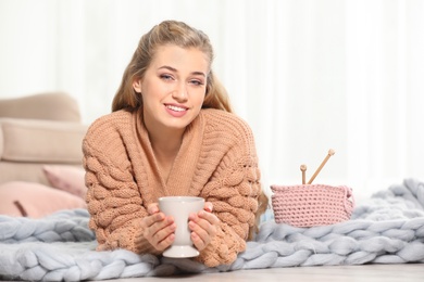 Photo of Attractive smiling young woman in cozy warm sweater with cup of hot drink lying on floor at home. Space for text