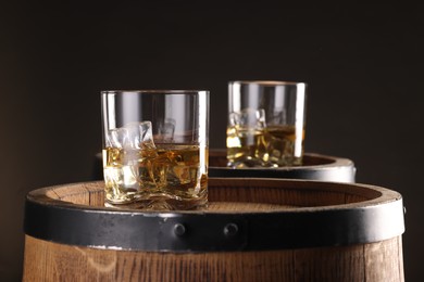 Whiskey with ice cubes in glasses on wooden barrels against dark background, closeup