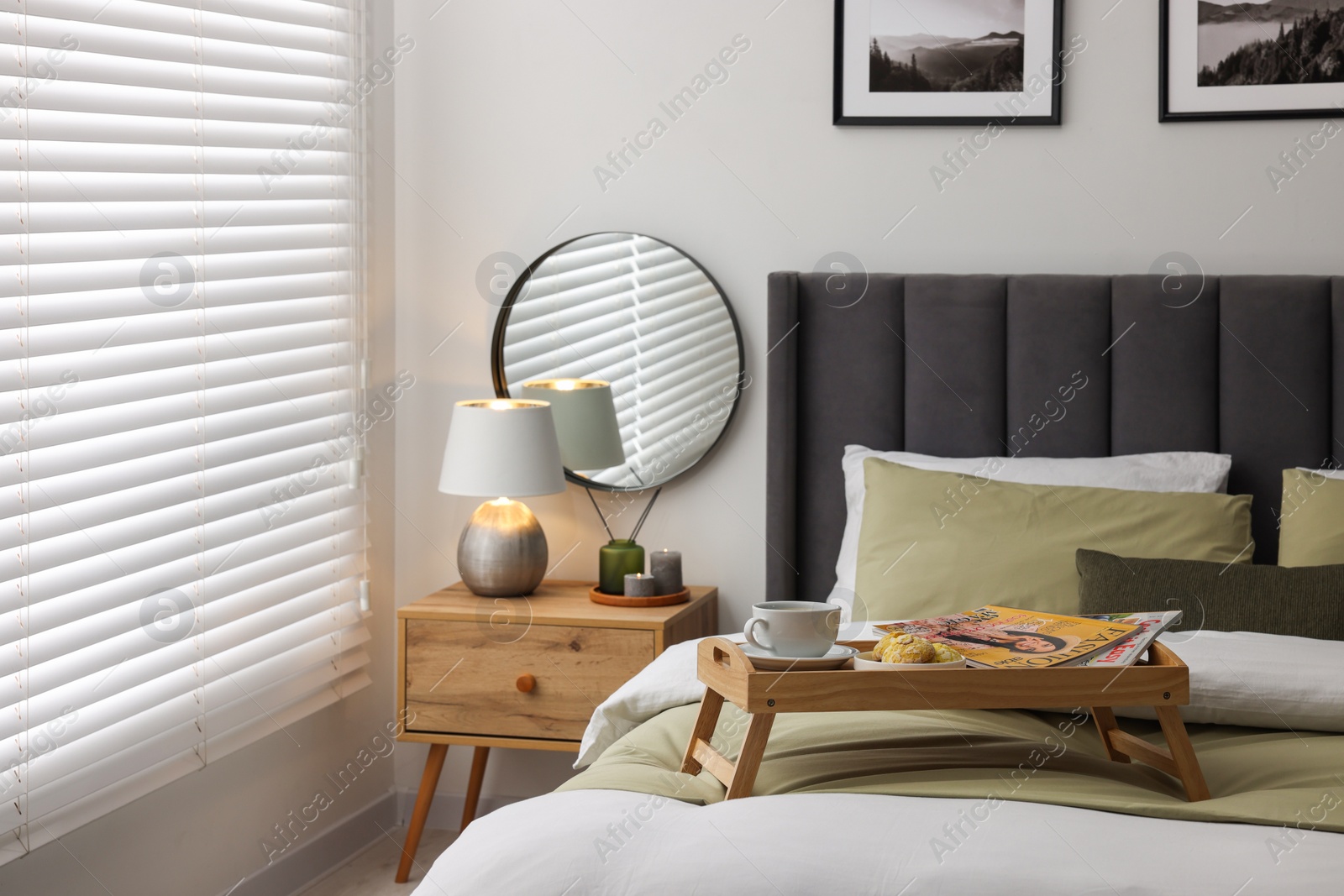 Photo of Wooden tray table with cup of drink and magazines on bed near window with horizontal blinds in room