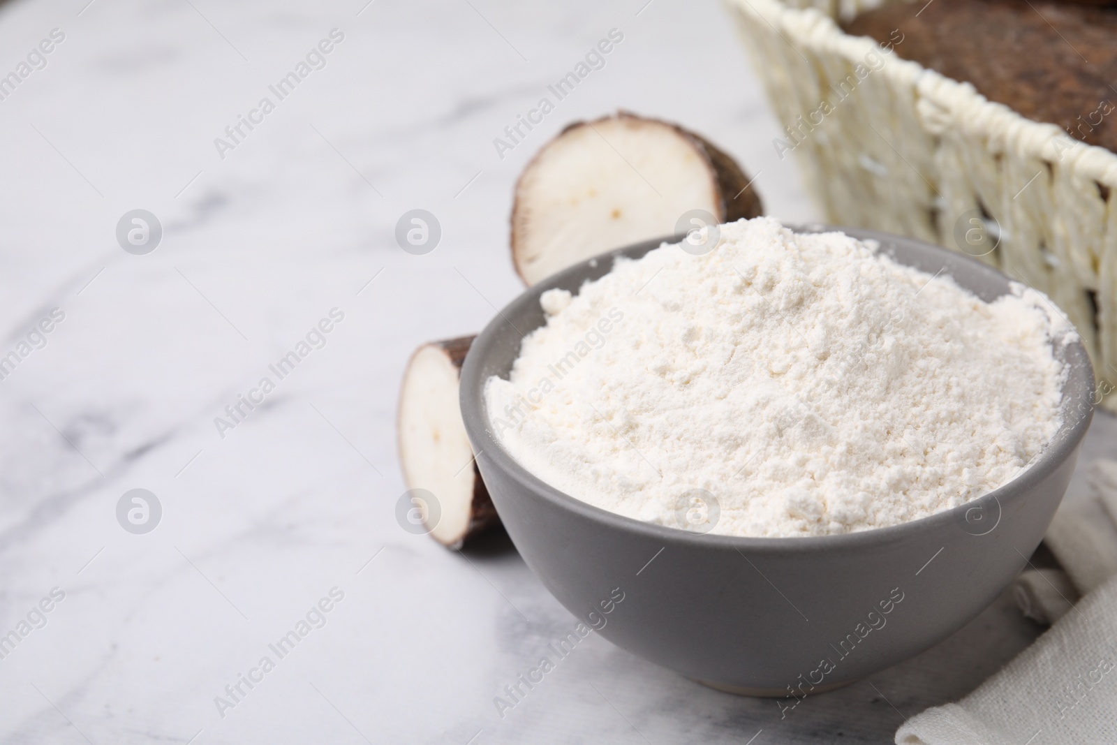 Photo of Bowl with cassava flour and roots on white marble table. Space for text