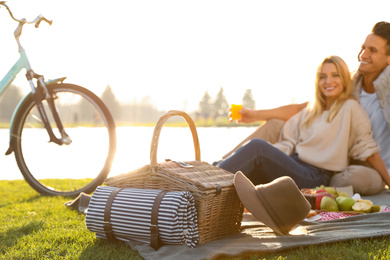 Photo of Happy young couple having picnic near lake, focus on wicker basket with blanket and hat