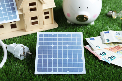 Photo of Solar panels, house model and money on green grass, closeup