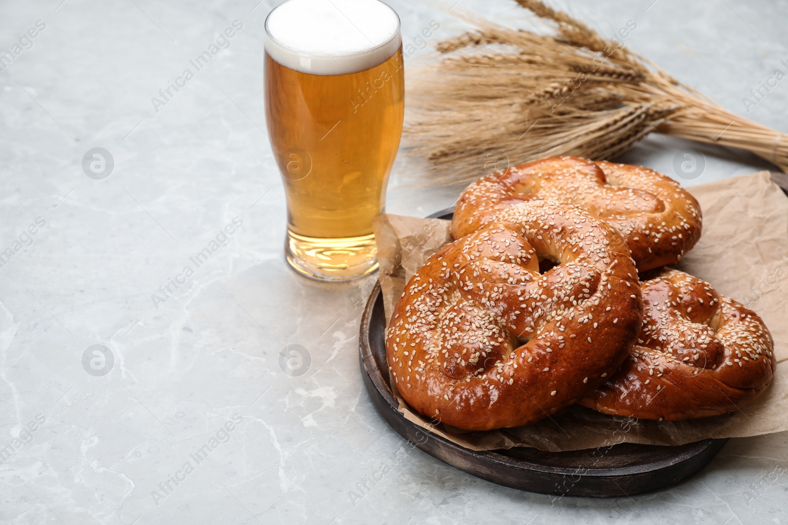 Photo of Tasty pretzels, glass of beer and wheat spikes on light grey table, space for text