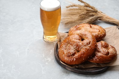 Tasty pretzels, glass of beer and wheat spikes on light grey table, space for text