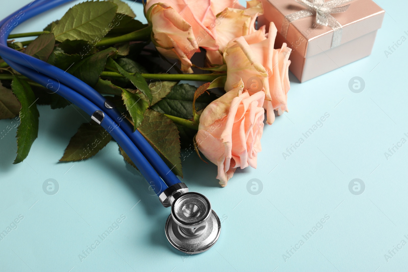 Photo of Stethoscope, gift box and flowers on light blue background, closeup with space for text. Happy Doctor's Day