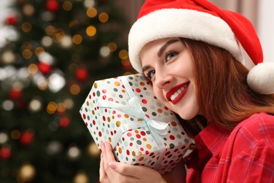 Photo of Beautiful young woman in Santa hat with gift box near Christmas tree indoors, space for text