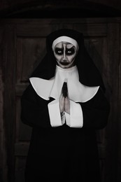 Photo of Portrait of scary devilish nun with clasped hands near wooden door outdoors. Halloween party look