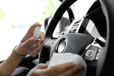 Photo of Woman cleaning steering wheel with wet wipe and antibacterial spray in car, closeup