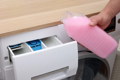 Photo of Man pouring fabric softener from bottle into washing machine indoors, closeup