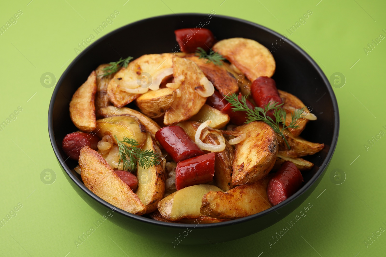 Photo of Delicious baked potato with thin dry smoked sausages, onion and dill in bowl on green table, closeup