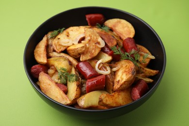 Photo of Delicious baked potato with thin dry smoked sausages, onion and dill in bowl on green table, closeup