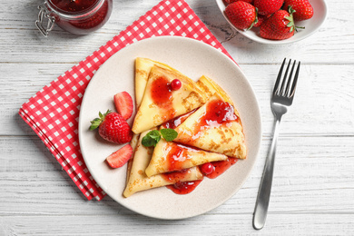 Photo of Delicious thin pancakes with strawberries and jam on white wooden table, flat lay