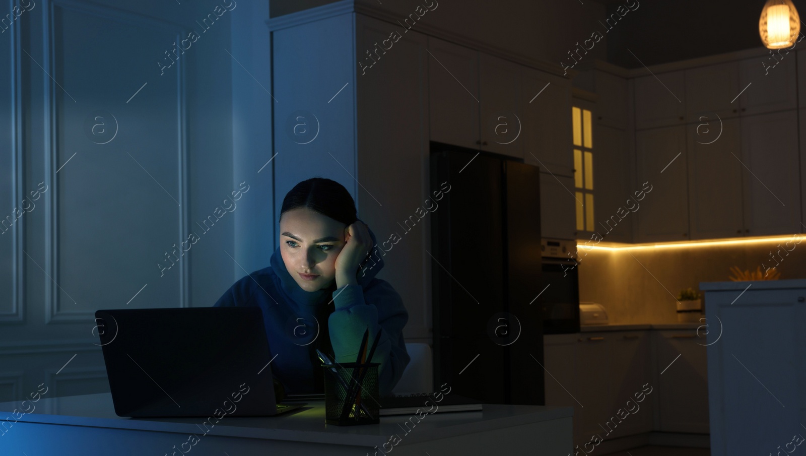 Photo of Young woman using laptop at table in kitchen at night