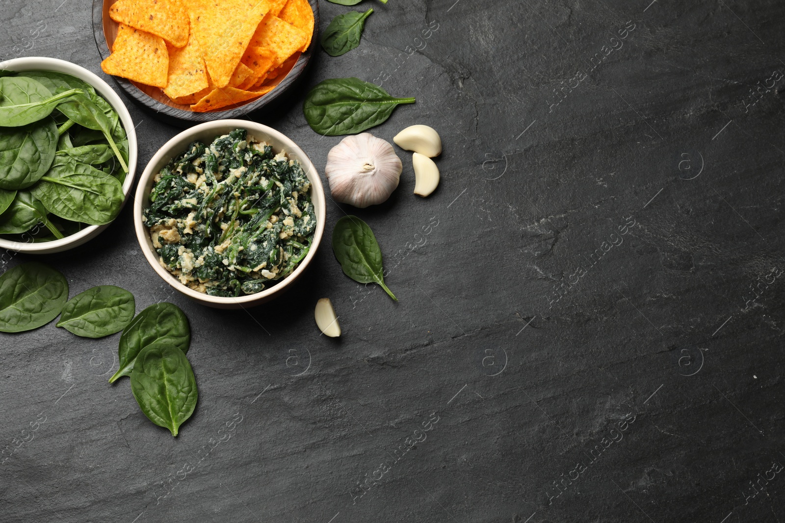 Photo of Tasty spinach dip with egg in bowl, garlic and nachos chips on black table, flat lay. Space for text