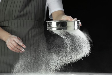 Woman sieving flour at table against black background, closeup