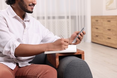 Photo of Man using smartphone while writing in notebook on sofa armrest wooden table at home, closeup