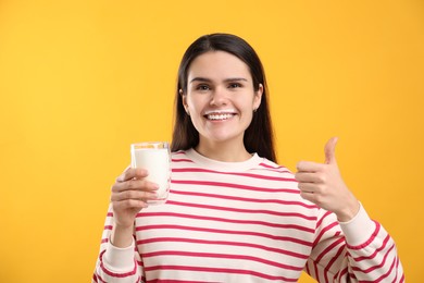Photo of Happy woman with milk mustache holding glass of tasty dairy drink and showing thumb up on yellow background